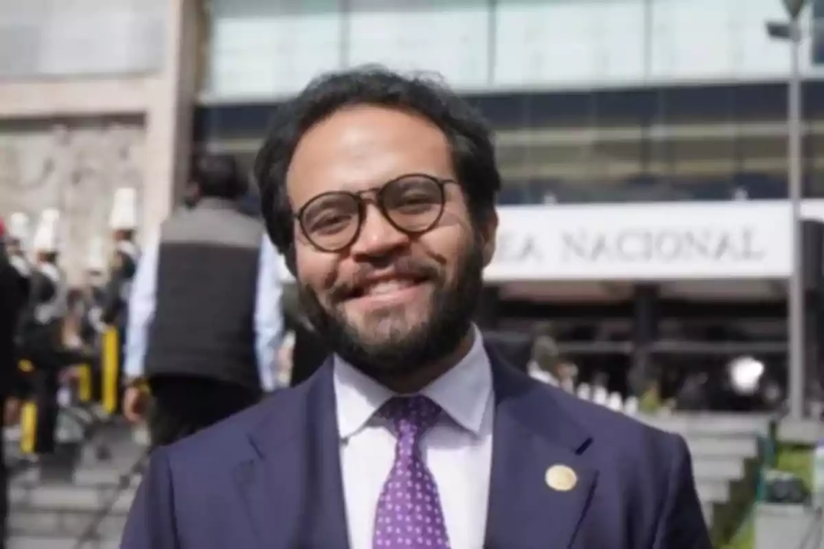 Man with a beard and glasses smiles while wearing a suit and purple tie in front of a public building.
