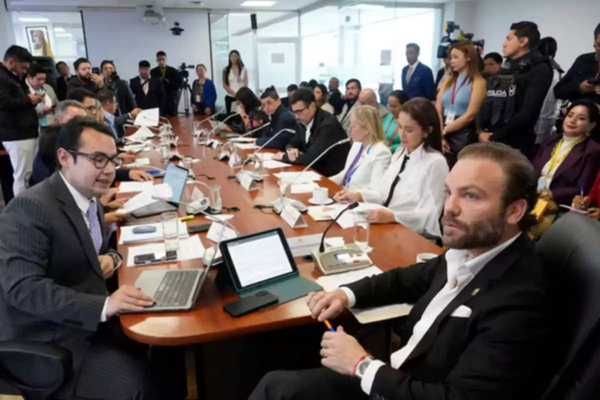 People gathered around a table in a conference room with laptops, documents, and microphones, while several others watch and take notes in the background.