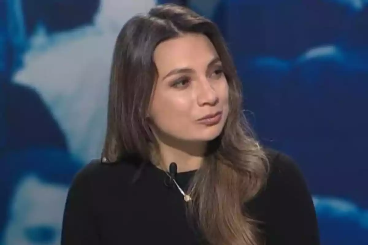 Long-haired brunette woman wearing a black sweater and a lapel microphone in a television studio with a blurred blue background