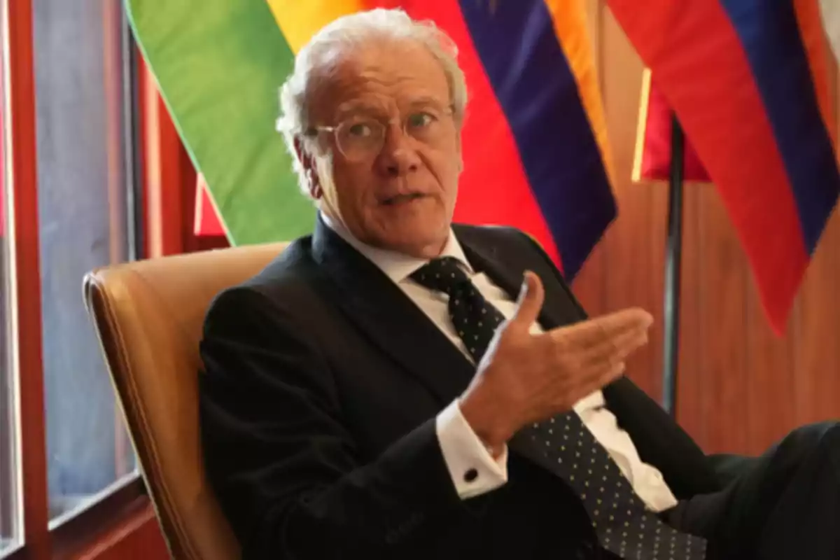 Elderly man with gray hair and glasses sitting in a chair, wearing a dark suit and tie, gesturing with his hand while speaking, with flags in the background.