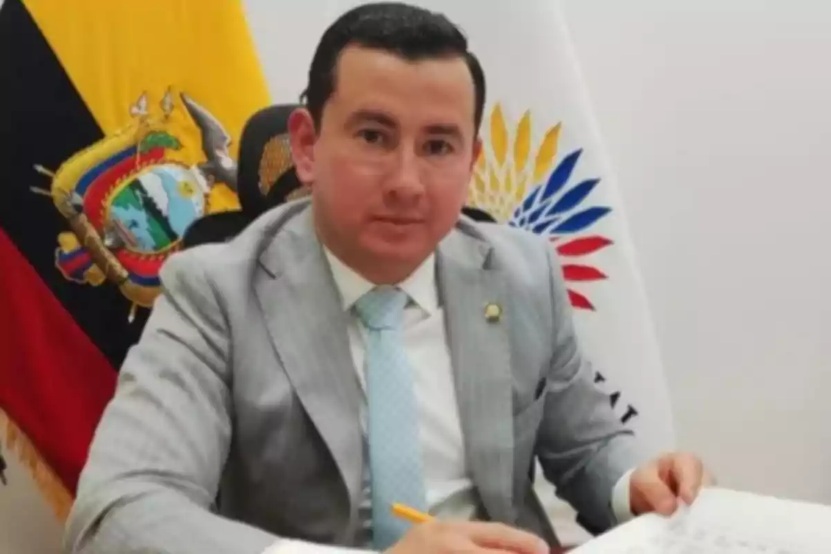 Man in a gray suit and light blue tie sitting at a desk with Ecuadorian flags in the background
