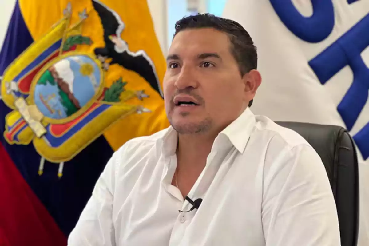 Hombre de cabello corto y camisa blanca sentado frente a una bandera de Ecuador y otra bandera blanca con letras azules