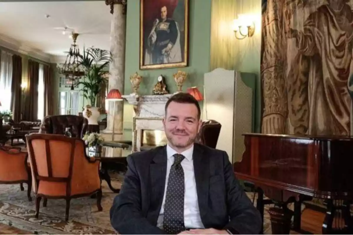 Man in a suit sitting in an elegant room with classic furniture, a piano, a fireplace, and an old portrait on the wall.