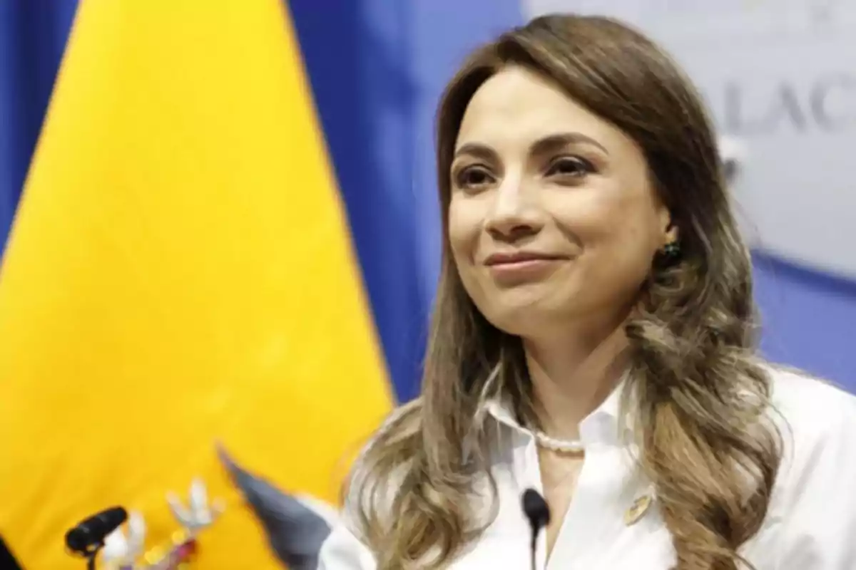 Light brown-haired woman with long hair, wearing a white jersey and a pearl necklace, speaking in front of a microphone with an Ecuadorian flag in the background.