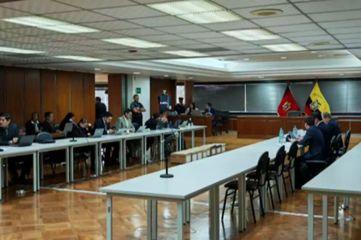 People gathered in a courtroom with official flags in the background and several white tables arranged in a U-shape.