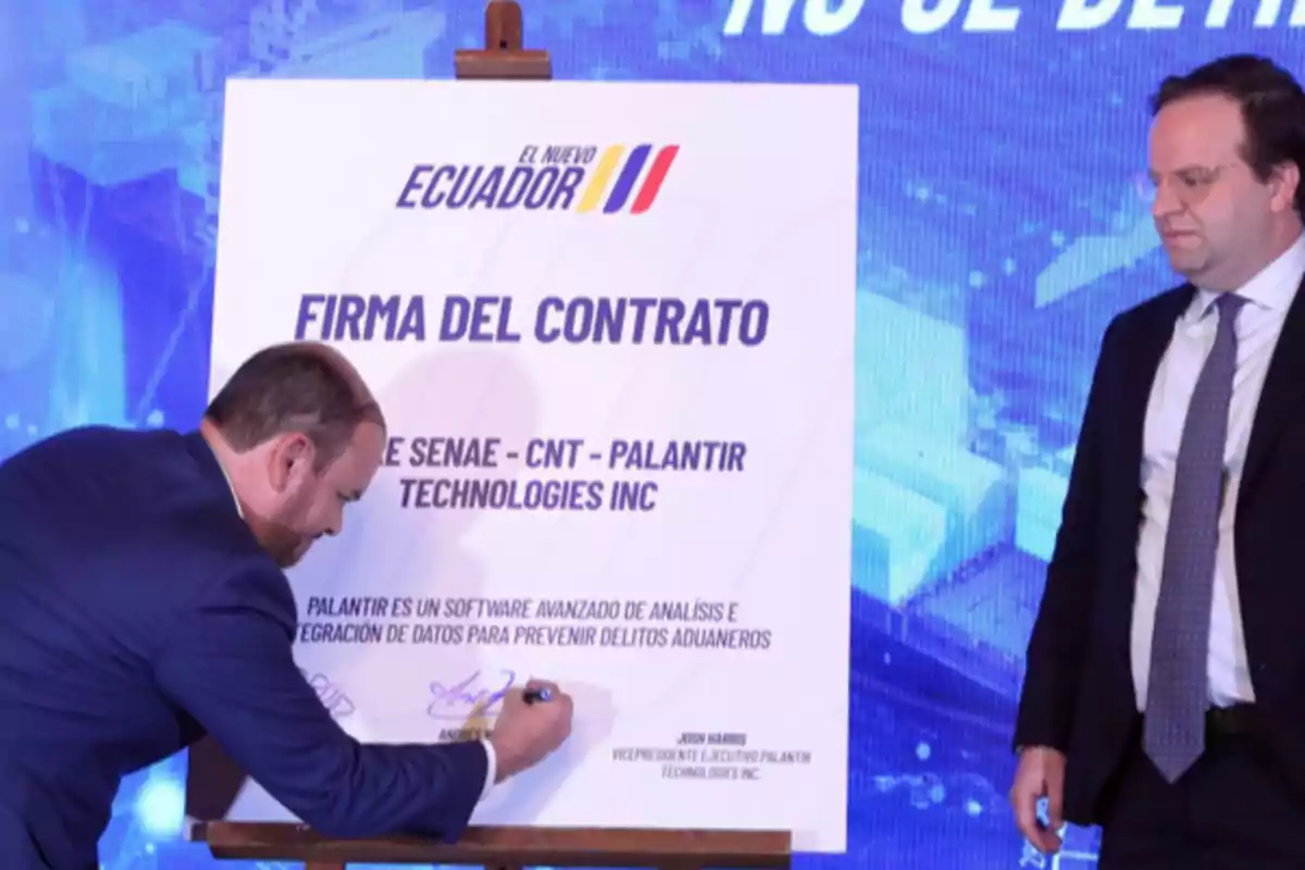A man signs a large poster announcing the signing of a contract between SENAE, CNT, and Palantir Technologies Inc while another man stands watching on the right.