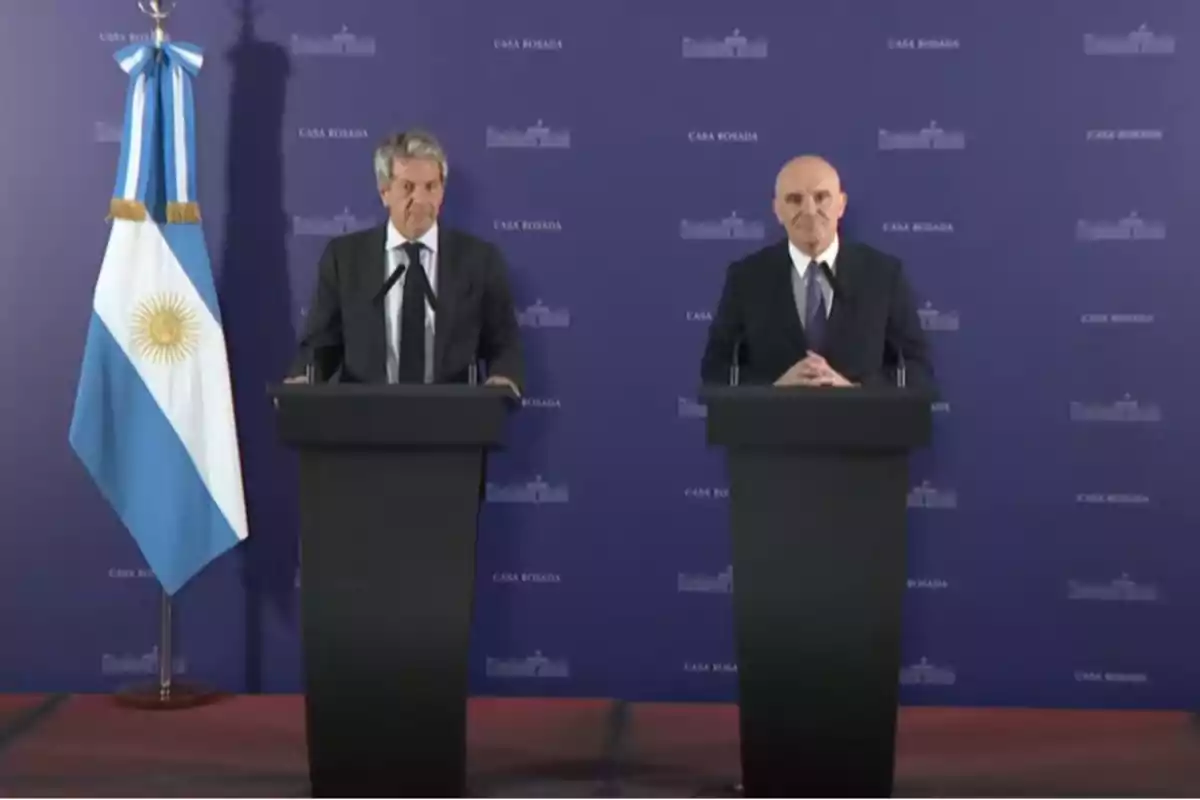 Two men in suits are standing behind black podiums at a press conference with a blue background and the Argentine flag on the left.