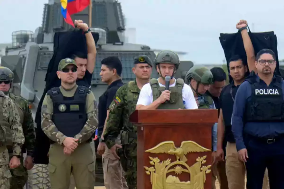 A group of military personnel and police officers escort a man wearing a helmet and a bulletproof vest who is speaking at a podium with the national emblem, while two handcuffed people raise their arms behind them and an armored vehicle is in the background.