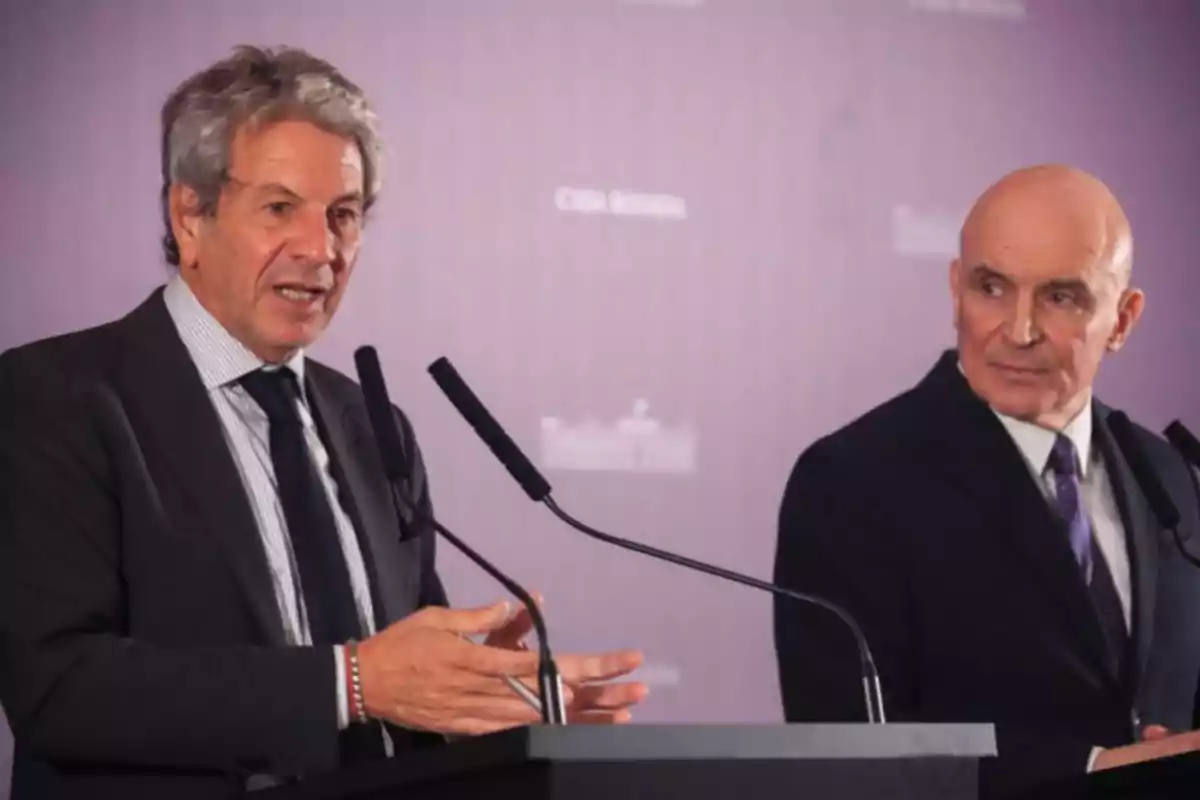 Dos hombres de traje hablando frente a micrófonos en un evento formal con fondo violeta.