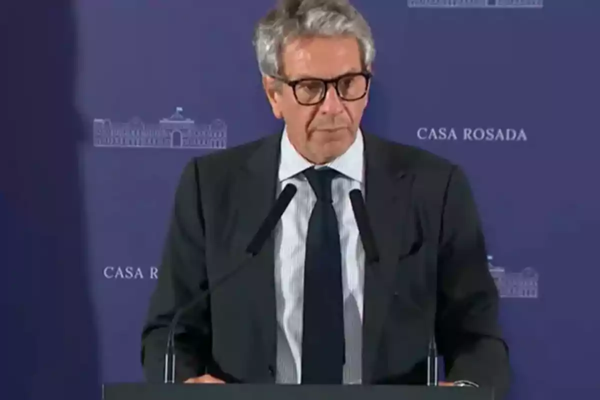 Gray-haired man with glasses speaking at a podium with a blue background that says Casa Rosada Gray-haired man with glasses speaking at a podium with a blue background that says Casa Rosada