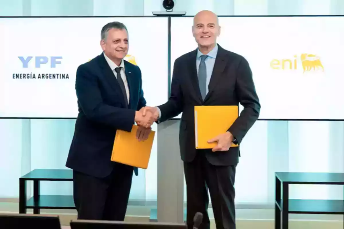 Dos hombres de traje se dan la mano mientras sostienen carpetas amarillas frente a pantallas con los logotipos de YPF y Eni Dos hombres de traje se dan la mano mientras sostienen carpetas amarillas frente a pantallas con los logotipos de YPF y Eni