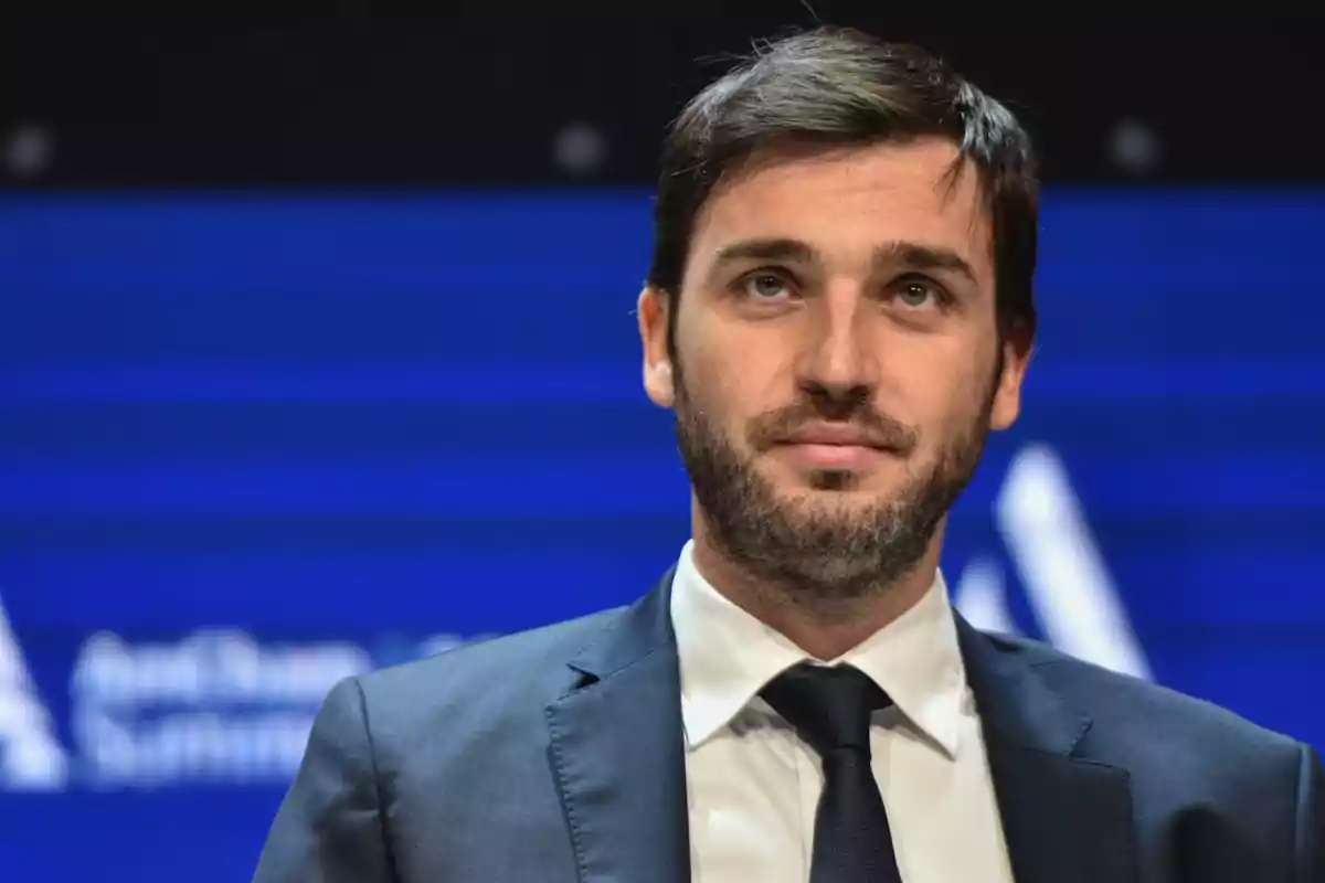 Hombre joven con barba y cabello corto usando traje y corbata frente a un fondo azul