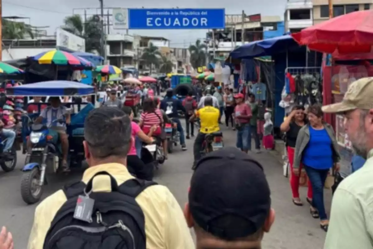 Personas caminando y circulando en motocicletas en una calle concurrida con puestos de mercado y un letrero azul que da la bienvenida a Ecuador Personas caminando y circulando en motocicletas en una calle concurrida con puestos de mercado y un letrero azul que da la bienvenida a Ecuador