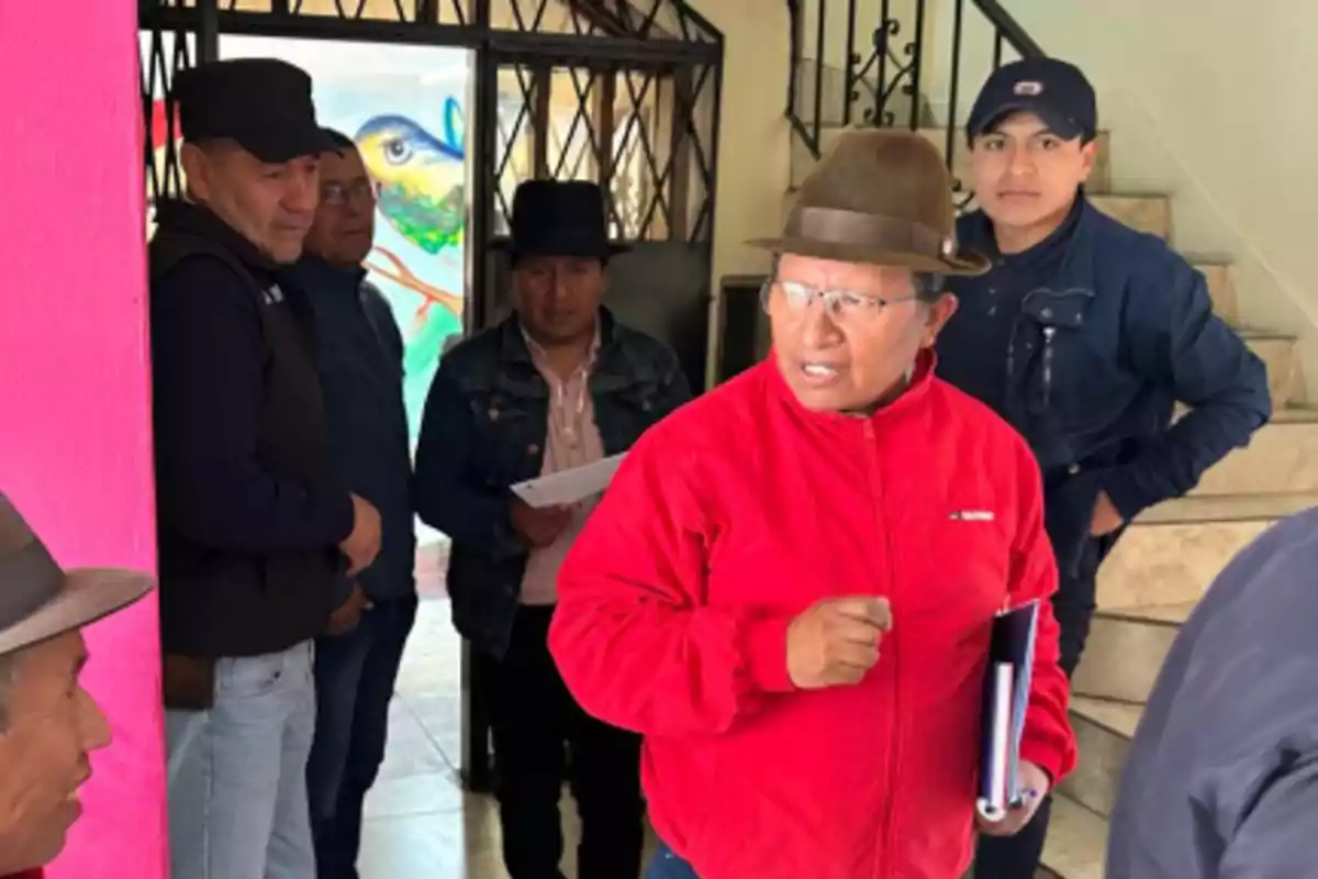 Un grupo de hombres con sombreros y gorras conversa en el interior de un edificio junto a una pared rosa y una escalera, uno de ellos lleva una chaqueta roja y sostiene documentos.