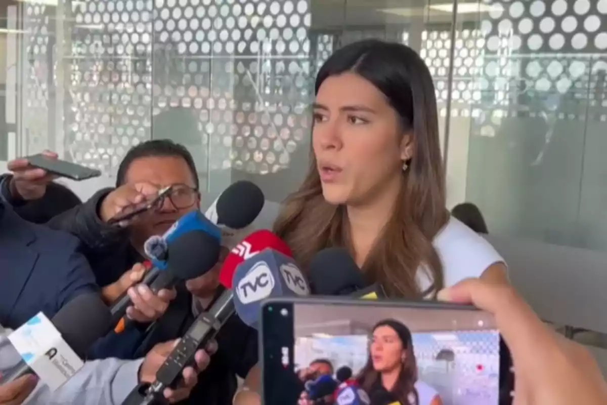 A young woman gives statements to several media outlets while holding an interview surrounded by microphones and recorders in an enclosed space with glass walls decorated with white circles. A young woman gives statements to several media outlets while holding an interview surrounded by microphones and recorders in an enclosed space with glass walls decorated with white circles.