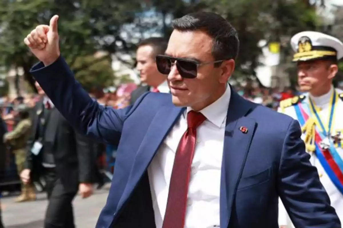 Man wearing sunglasses and a blue suit giving a thumbs-up at an outdoor public event