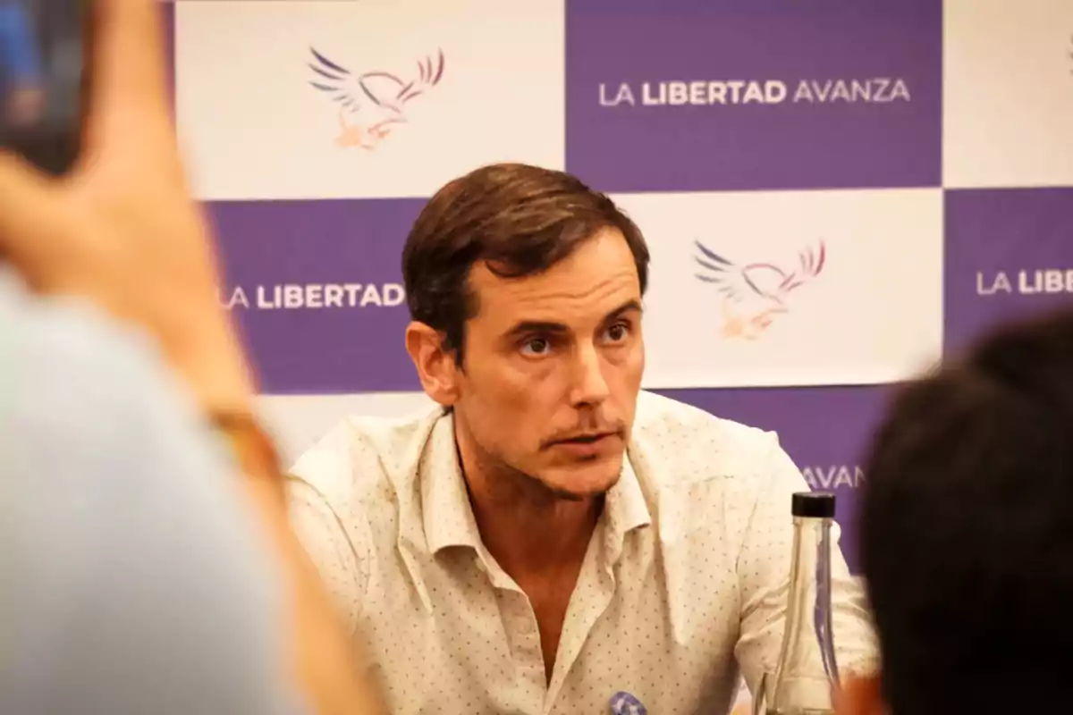 Hombre de cabello corto y camisa clara sentado frente a un fondo con el logo y el texto La Libertad Avanza mientras es fotografiado por varias personas