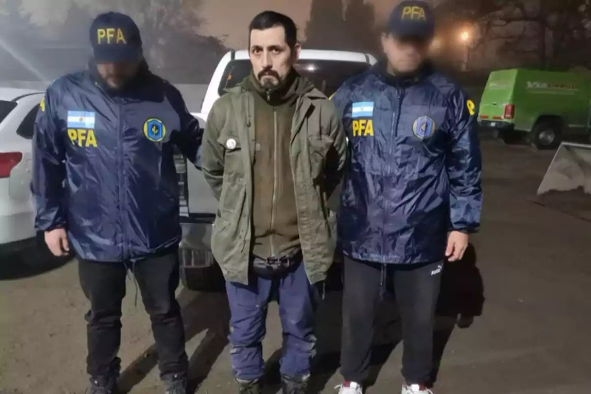 Tres hombres de pie, uno en el centro con las manos hacia atrás y dos a los costados con chaquetas azules y gorras con las siglas PFA, en un entorno exterior nocturno.