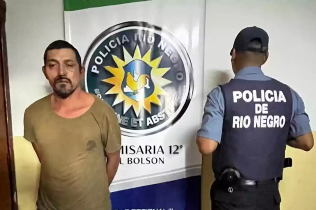 Un hombre de camiseta marrón está de pie junto a un policía de espaldas con chaleco que dice Policía de Río Negro frente a un cartel de la comisaría Un hombre de camiseta marrón está de pie junto a un policía de espaldas con chaleco que dice Policía de Río Negro frente a un cartel de la comisaría