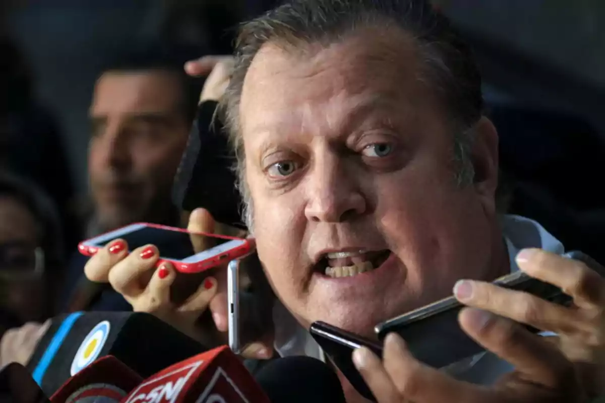 Hombre hablando con la prensa rodeado de micrófonos y teléfonos móviles
