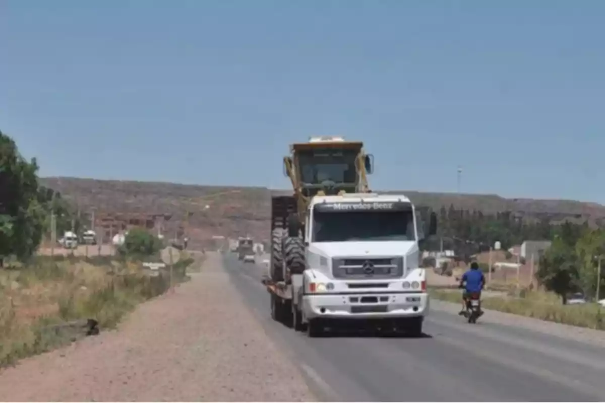 Camión blanco transportando maquinaria pesada por una carretera rural bajo un cielo despejado