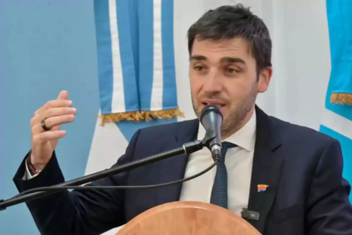 Hombre de traje hablando en un micrófono con banderas celestes y blancas de fondo Hombre de traje hablando en un micrófono con banderas celestes y blancas de fondo