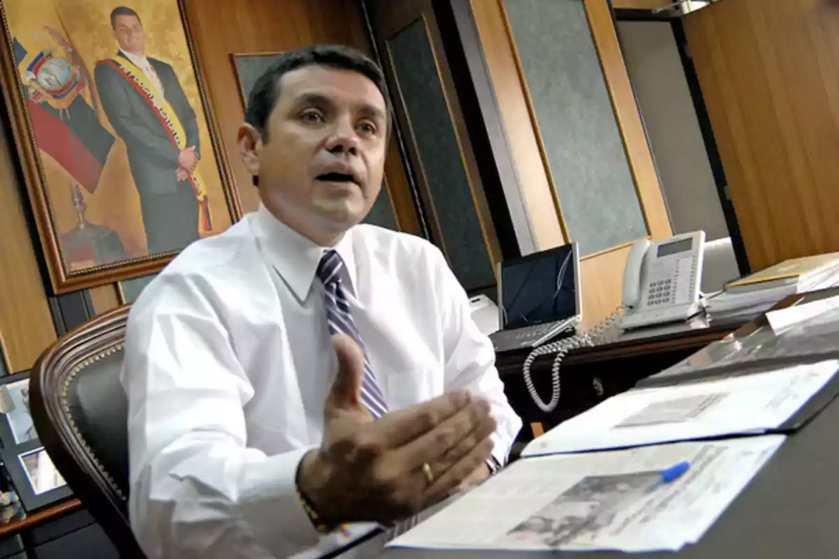 Man in a suit and tie sitting in an office talking, with documents on the desk and an official portrait on the wall behind him
