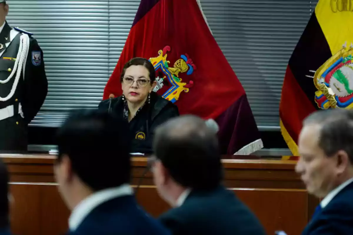 Una jueza sentada en una sala de audiencias con banderas oficiales detrás y varias personas en primer plano Una jueza sentada en una sala de audiencias con banderas oficiales detrás y varias personas en primer plano