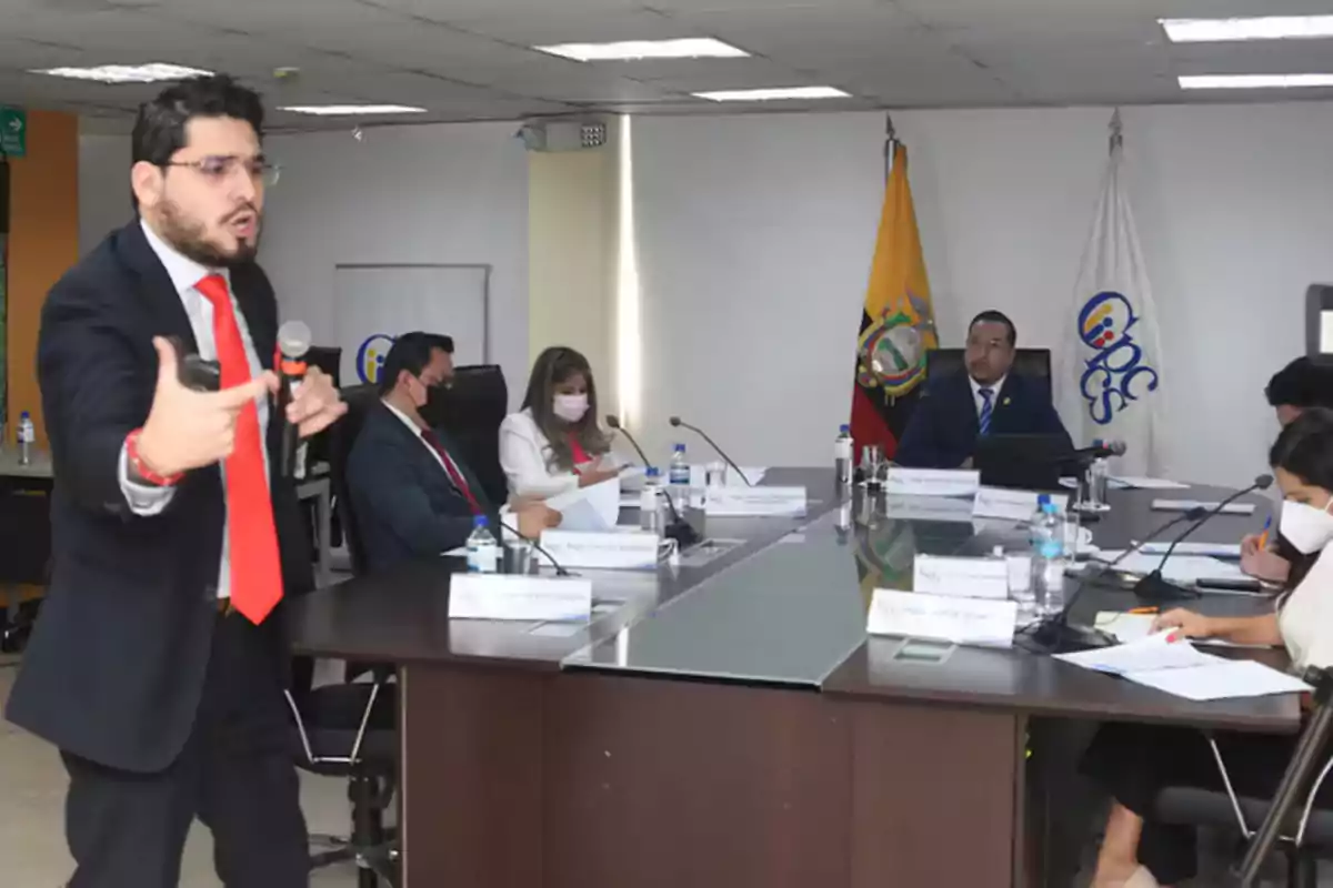 Un hombre de traje y corbata roja habla frente a una mesa de reunión donde varias personas están sentadas tomando notas y usando mascarillas, al fondo hay banderas y un logotipo institucional.