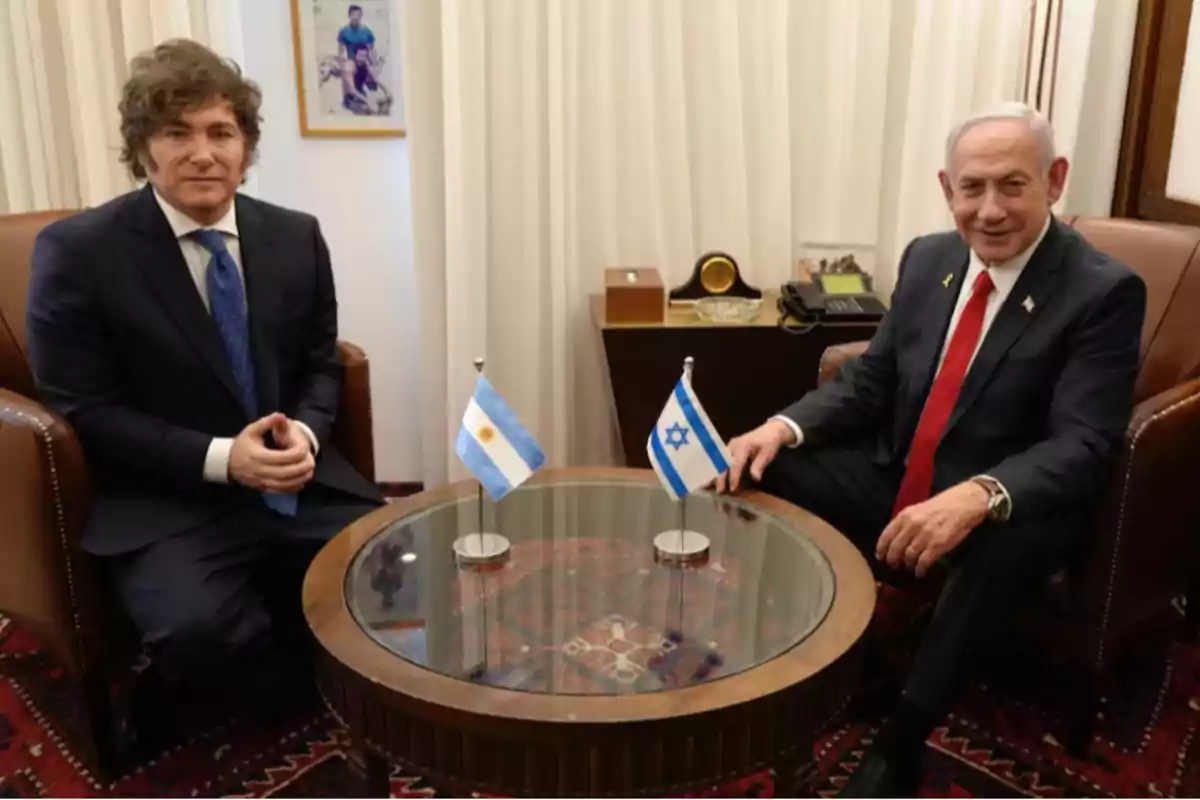Dos hombres con traje sentados en sillones frente a una mesa redonda de vidrio con las banderas de Argentina e Israel