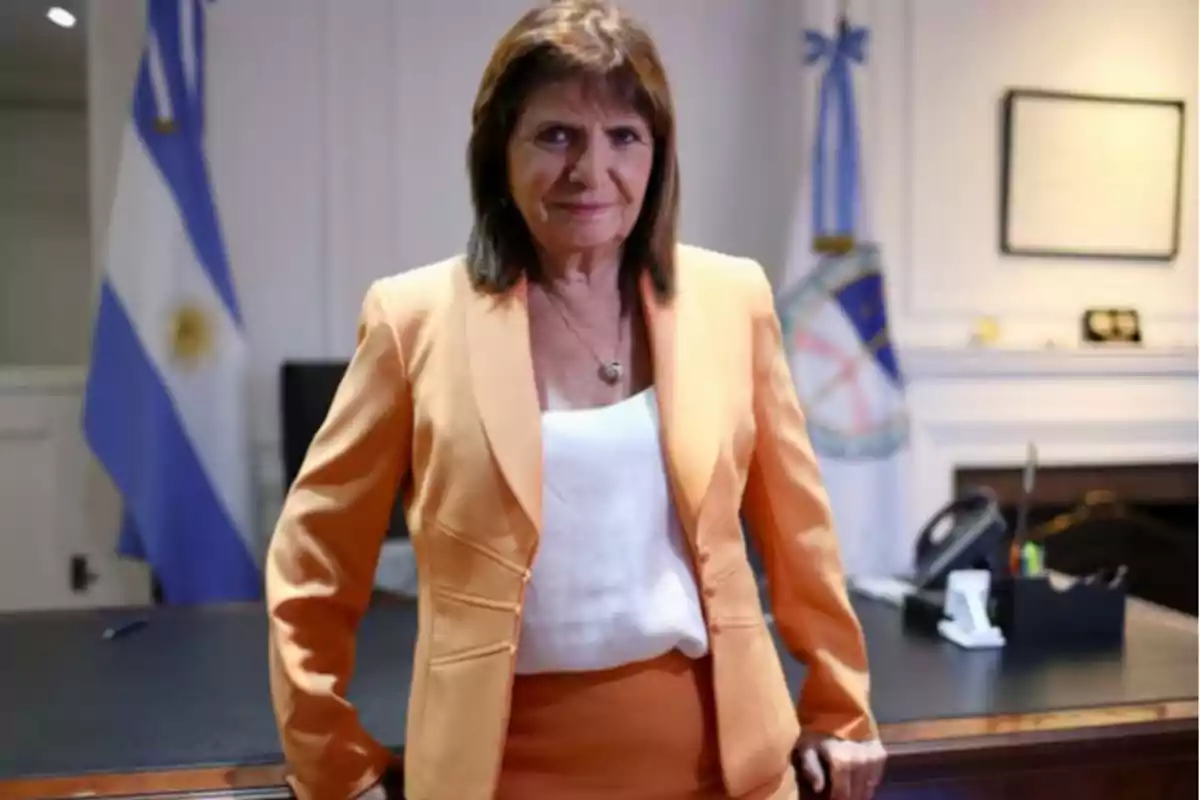 Mujer de cabello castaño con saco naranja y blusa blanca de pie frente a un escritorio en una oficina con banderas argentinas al fondo