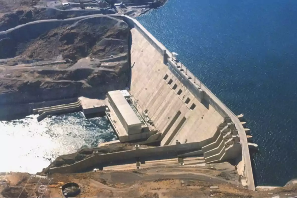 Vista aérea de una represa de concreto junto a un embalse de agua rodeada de montañas y caminos.