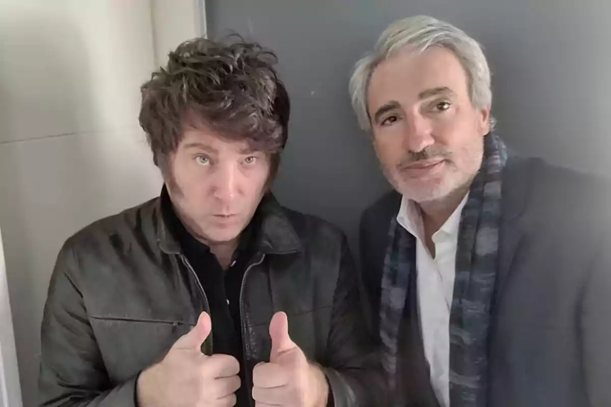 Dos hombres posan juntos, uno de ellos con chaqueta oscura hace gesto de pulgares arriba y el otro lleva saco y bufanda