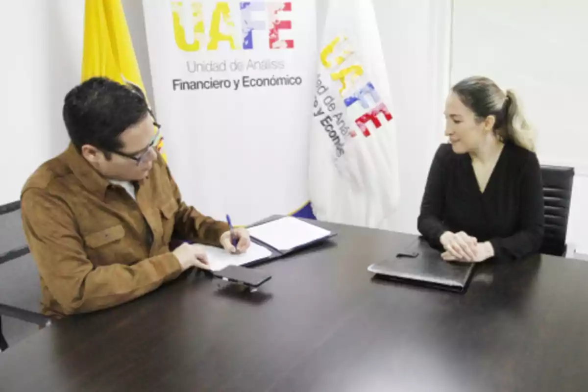 Two people sitting at an office table while one of them signs a document and the other watches; in the background, a yellow flag and a sign with the UAFE logo can be seen.