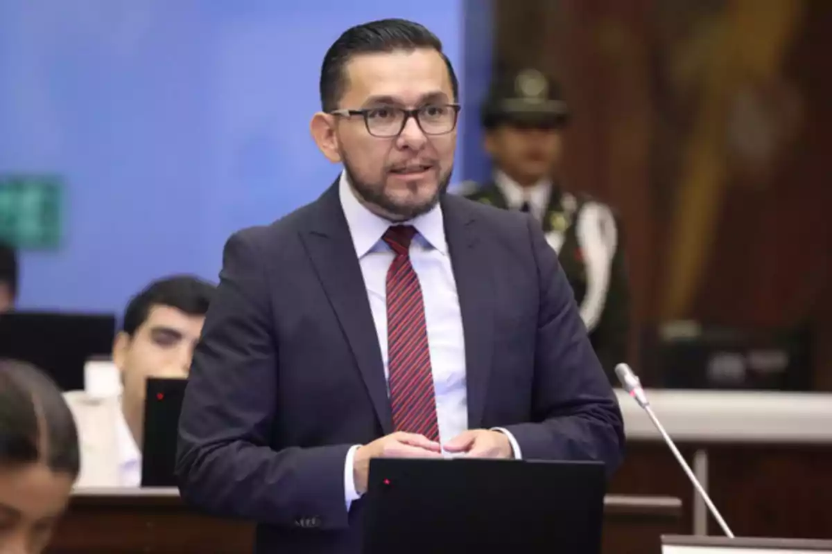 Man with glasses and a beard wearing a suit and red tie standing in front of a microphone in a formal setting with people and computers in the background