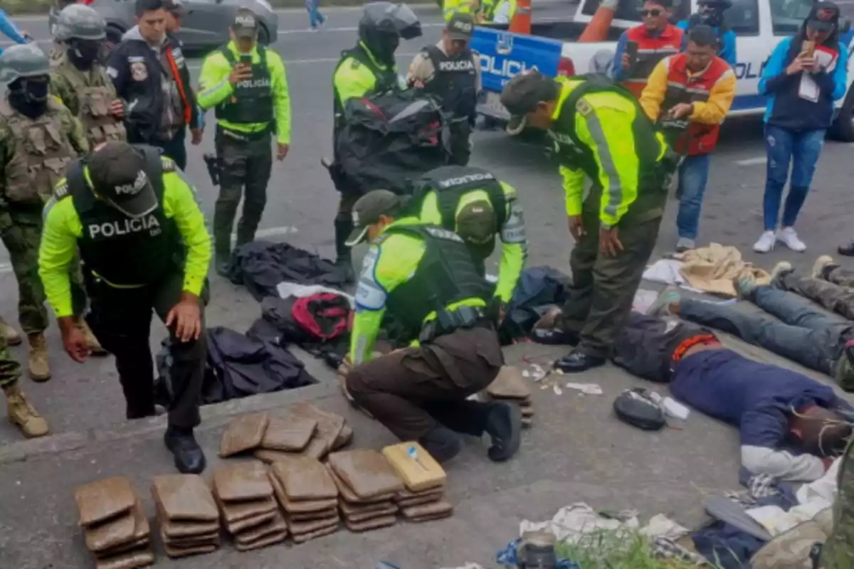 Police inspect drug packages on the ground while several people are detained and lying face down on a street surrounded by officers and journalists.