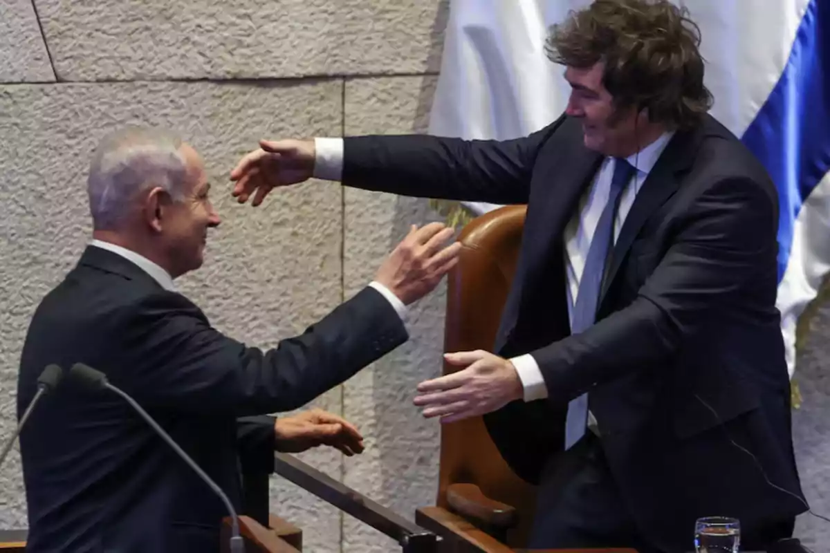Dos hombres de traje se saludan sonrientes y se disponen a abrazarse en un entorno formal con banderas de fondo. Dos hombres de traje se saludan sonrientes y se disponen a abrazarse en un entorno formal con banderas de fondo.