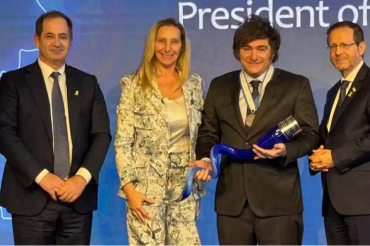 Cuatro personas vestidas de manera formal posan para una foto, una de ellas sostiene un objeto azul en forma de cuerno y lleva una medalla. Cuatro personas vestidas de manera formal posan para una foto, una de ellas sostiene un objeto azul en forma de cuerno y lleva una medalla.