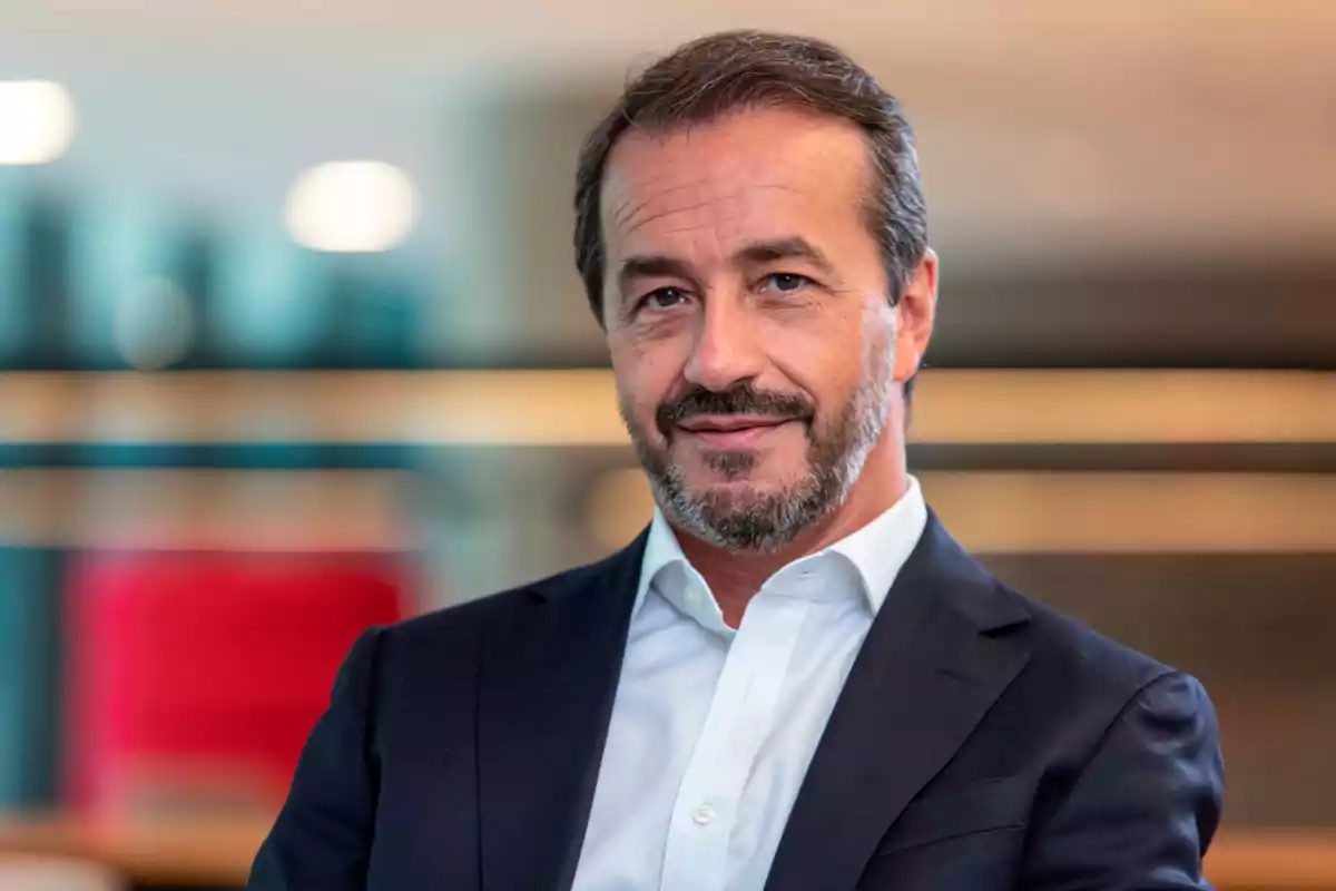 Hombre de mediana edad con barba y cabello corto, vestido con traje oscuro y camisa blanca, sonriendo levemente en un entorno profesional desenfocado. Hombre de mediana edad con barba y cabello corto, vestido con traje oscuro y camisa blanca, sonriendo levemente en un entorno profesional desenfocado.