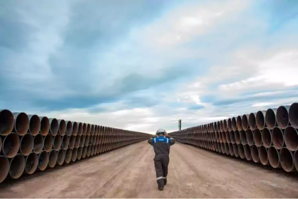 Worker walking between rows of large pipes stacked under a cloudy sky