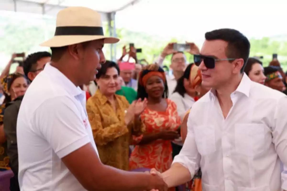 Dos hombres se dan la mano en un evento al aire libre mientras varias personas observan y aplauden en el fondo