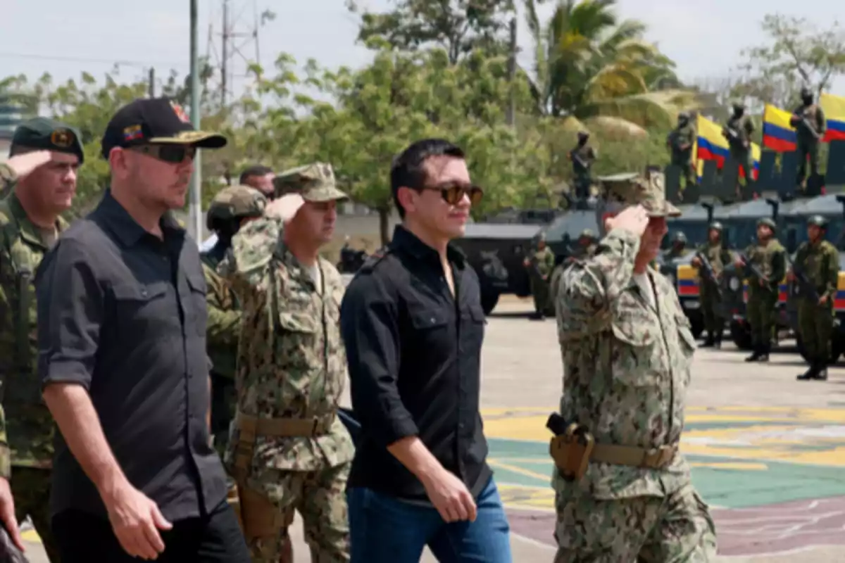 Varios militares y un civil caminan juntos en una base militar mientras algunos soldados saludan y al fondo se ven vehículos blindados y banderas de Colombia Varios militares y un civil caminan juntos en una base militar mientras algunos soldados saludan y al fondo se ven vehículos blindados y banderas de Colombia