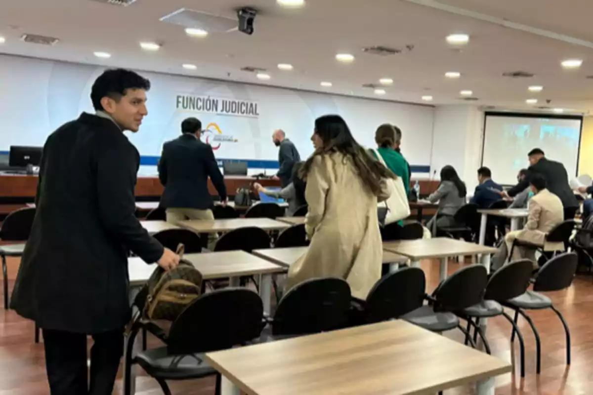 People in a courtroom with desks and chairs, some standing and others sitting, while in the background there is a projector and a sign that says "judicial function."