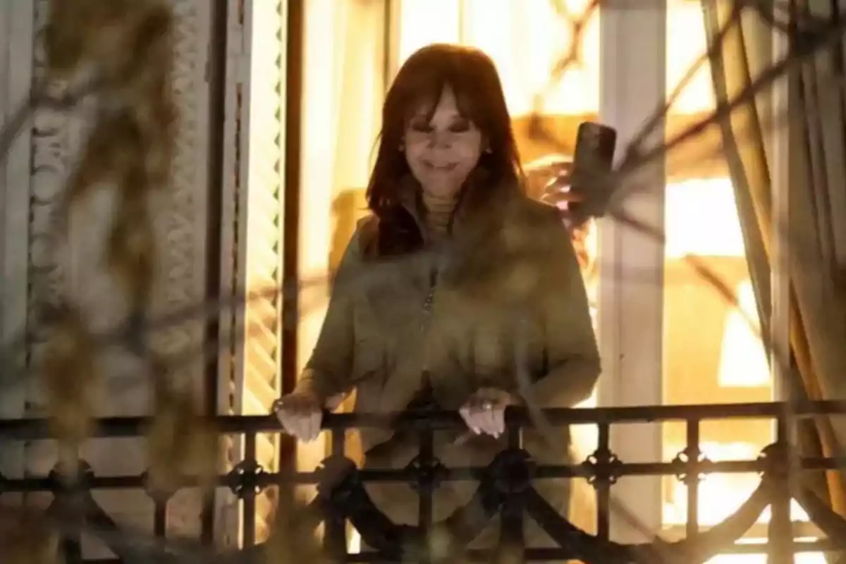 Brown-haired woman smiles while leaning on the railing of a balcony lit by the interior light of a house