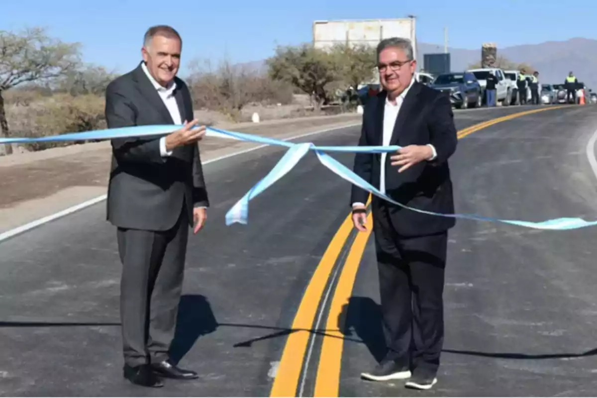 Two men in suits cut a light blue and white ribbon at the opening of a new road.