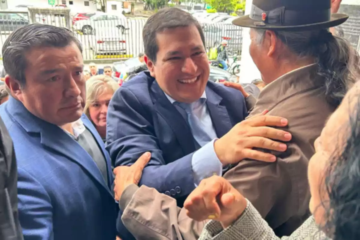 A smiling man in a suit greets and hugs another person wearing a hat in an outdoor setting surrounded by several people.