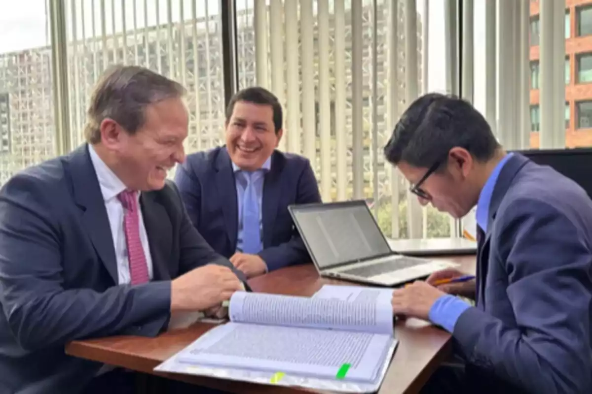 Three men in suits smile and talk while reviewing documents and a laptop in an office with large windows. Three men in suits smile and talk while reviewing documents and a laptop in an office with large windows.