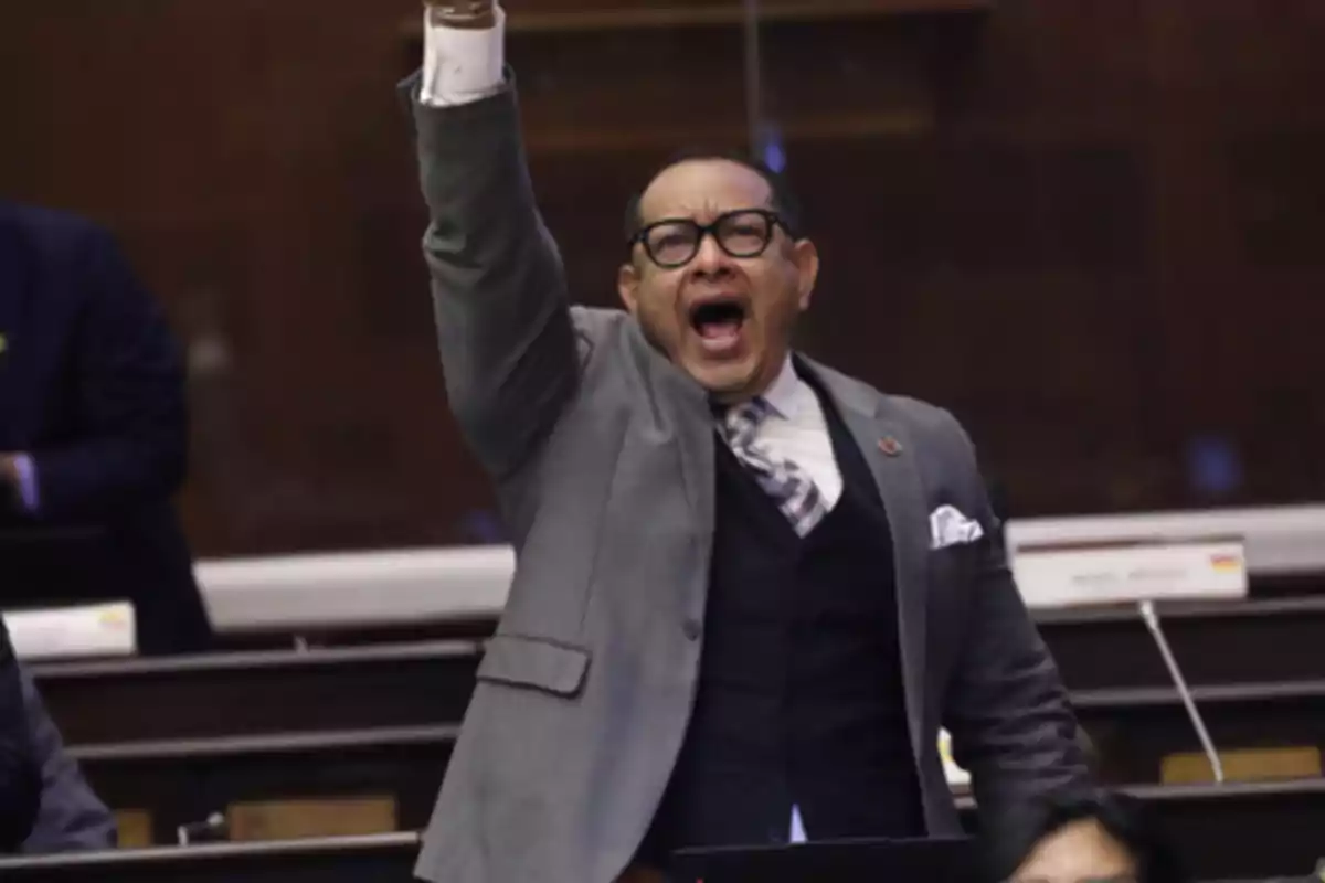 Man in a suit and glasses raising his arm and shouting in what appears to be a parliamentary chamber