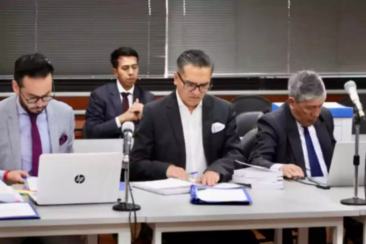 Four men in suits and ties are seated at a work table with laptops, microphones, and documents, in what appears to be a meeting room or courtroom.