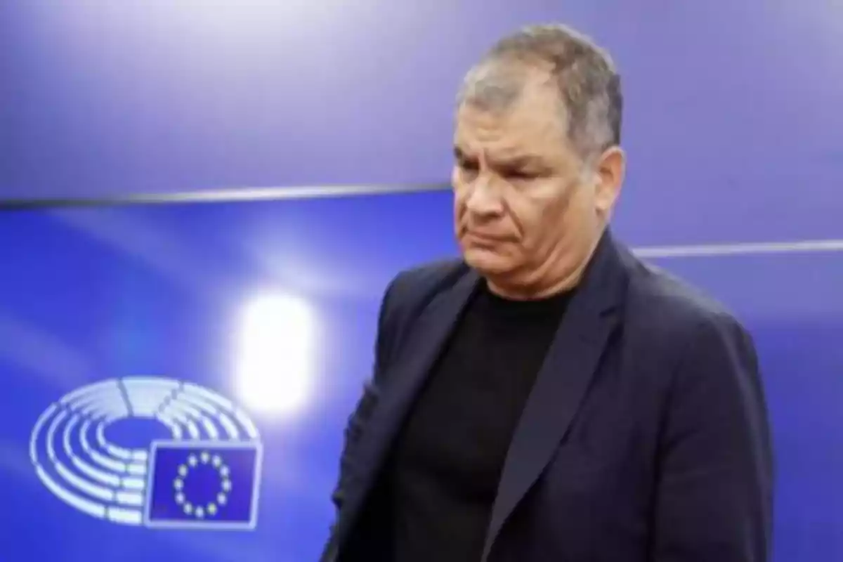 Gray-haired man with a serious expression wearing a dark jacket in front of a blue background with the European Union logo Gray-haired man with a serious expression wearing a dark jacket in front of a blue background with the European Union logo